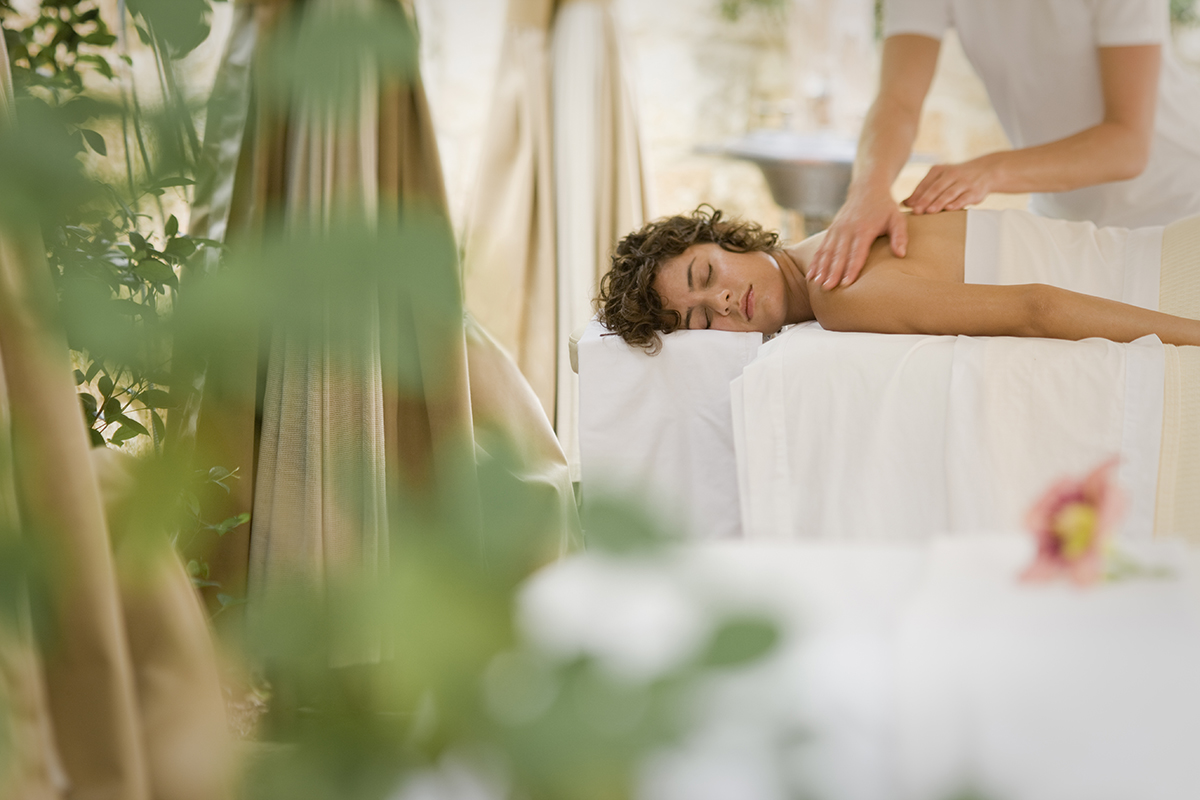 An outdoor spa treatment area surrounded by lush greenery and trees. There is a comfortable massage table covered with white sheets and decorated with a flower. A therapist is standing next to the table providing a relaxing treatment.