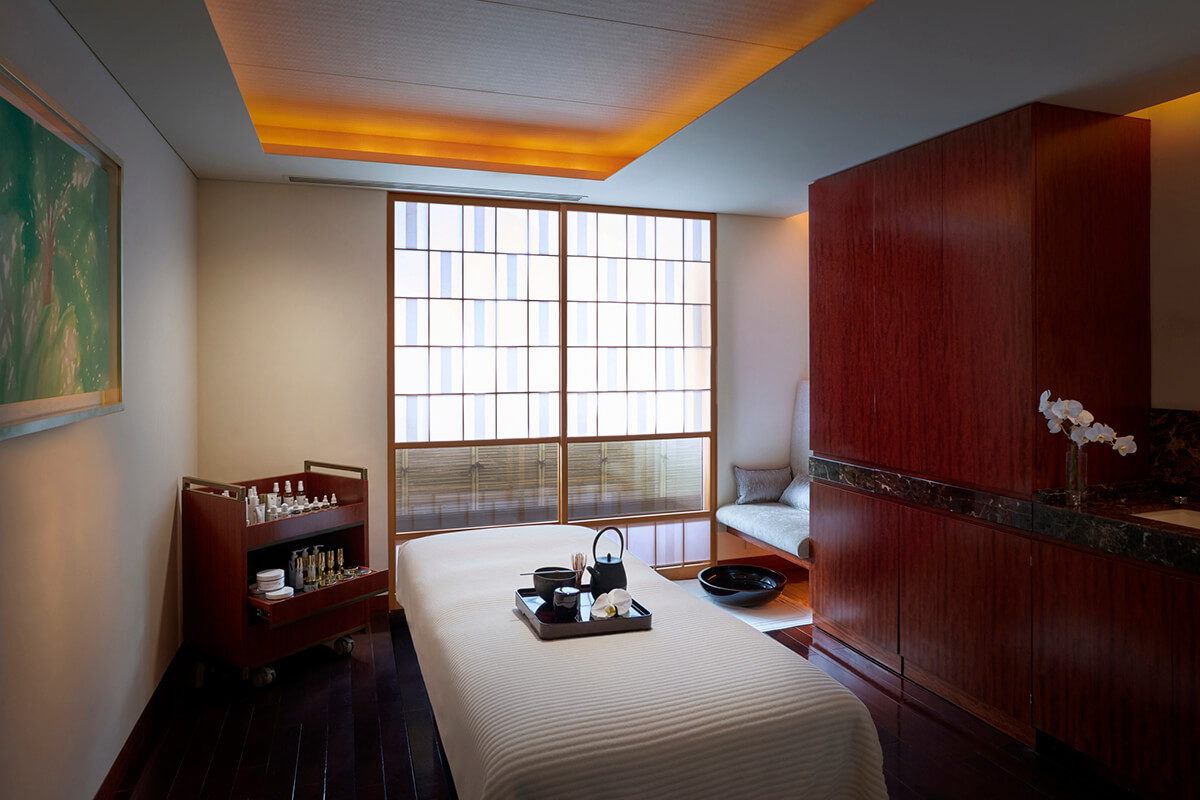 A serene and inviting treatment room at the Peninsula Tokyo spa, featuring a massage bed draped in white linens and surrounded by light-colored wooden panels. 