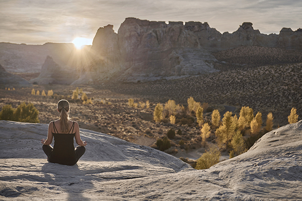 A person overlooking a vast, arid desert landscape wearing black yoga pants and a black tank top. She has her arms on her thighs in a meditation pose. The rocky ledge is high up and offers a breathtaking view of the desert valley below, with mountains in the distance at Amangiri in Utah. 