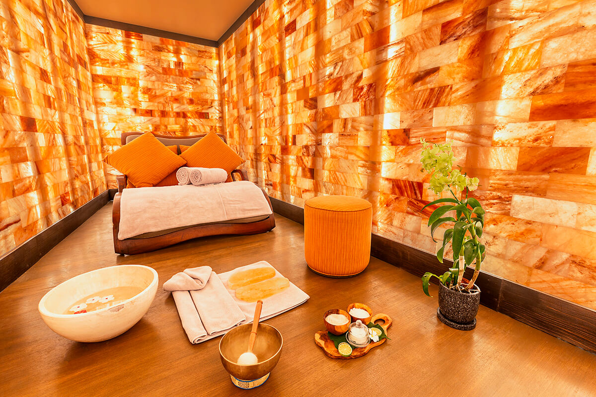 The interior of a salt room in a spa. The room has a serene and calming ambiance with dim lighting and walls made of salt bricks. There are several illuminated salt crystals on display. In the center of the room, there is a comfortable reclining chair with a blanket and a pillow.