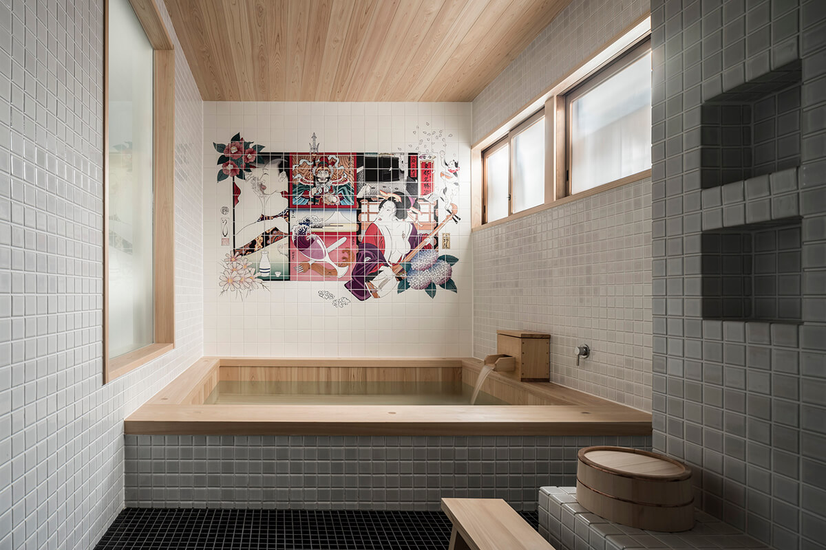 A traditional Japanese-style room in the Kakurenbo Yokocho Kagurazaka Trunk House, a luxury ryokan in Japan. The room features tiled floors and walls with an intricicate arwork of a Geisha. There is a bath in the center with running water with wooden seating.