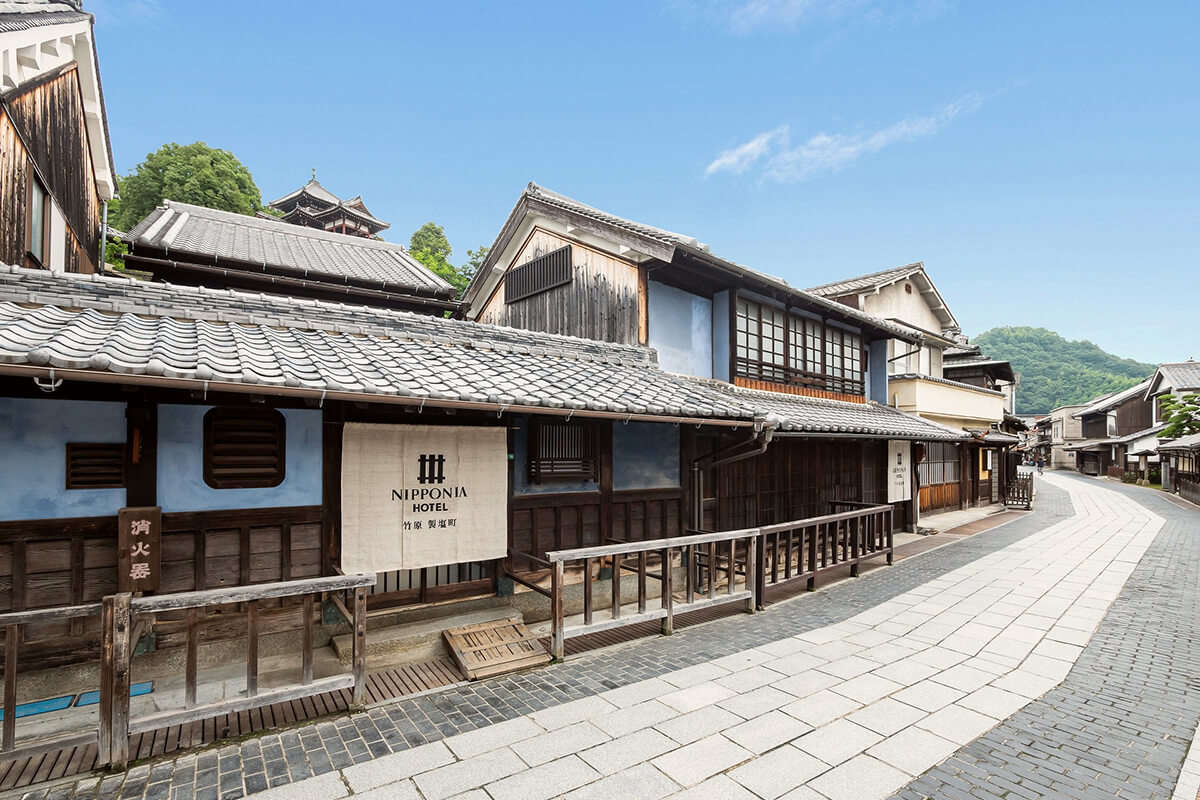 A traditional Japanese-style building nestled among trees with a small garden in front. The building has a tiled roof and wooden walls with large windows. In the foreground, there is a wooden deck with a table and chairs, and a few potted plants. Beyond the building, a misty forest can be seen, and in the background, there are mountains. The image captures the serene beauty of the natural surroundings and the traditional Japanese architecture.