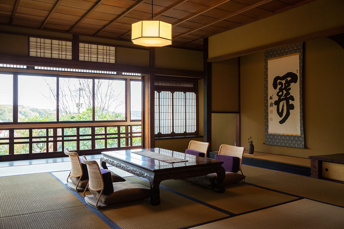 A traditional Japanese-style room in a Ryokan, or traditional Japanese inn, with tatami mats covering the floor and a low wooden table and cushions for seating. The room features sliding shoji doors and a tokonoma, a small alcove in the wall used for displaying artwork or flower arrangements. The overall ambiance of the room is tranquil and serene, providing a peaceful retreat for travelers seeking a traditional Japanese experience.