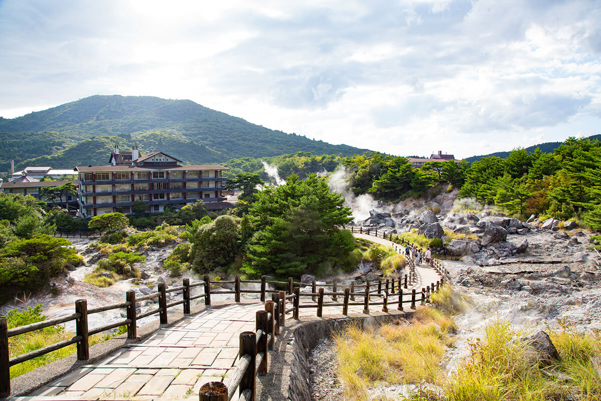 A serene outdoor scene showcasing a traditional Japanese hot spring, known as an onsen, located in Unzen, Japan. The image features a partially enclosed wooden structure that houses the hot spring, with steam rising from the water's surface. The onsen is surrounded by trees, foliage, and rocky terrain, creating a tranquil and natural setting. In the foreground, a small wooden bench can be seen where visitors can sit and soak in the hot spring while enjoying the beautiful view.
