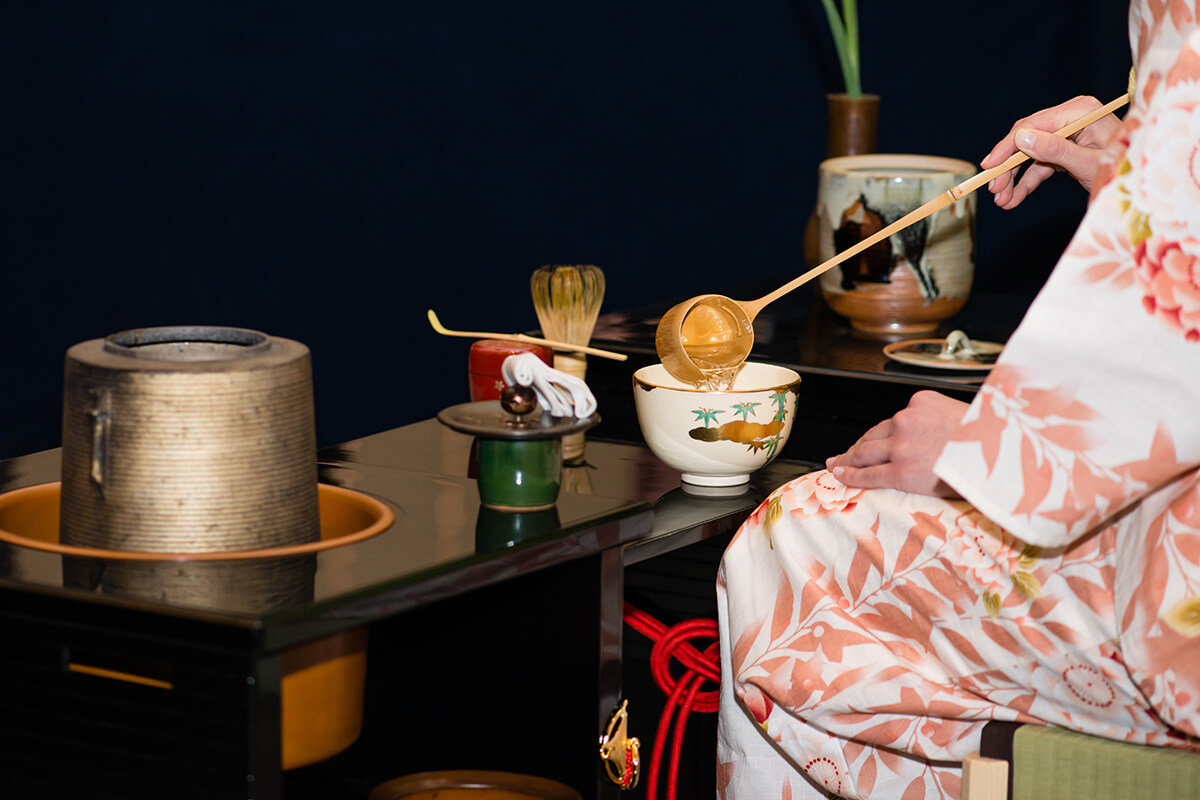 A young Japanese woman dressed in a traditional kimono is seen performing a tea ceremony in a serene and minimalist setting. There is a small table in front of her that holds a ceramic bowl and a bamboo whisk.