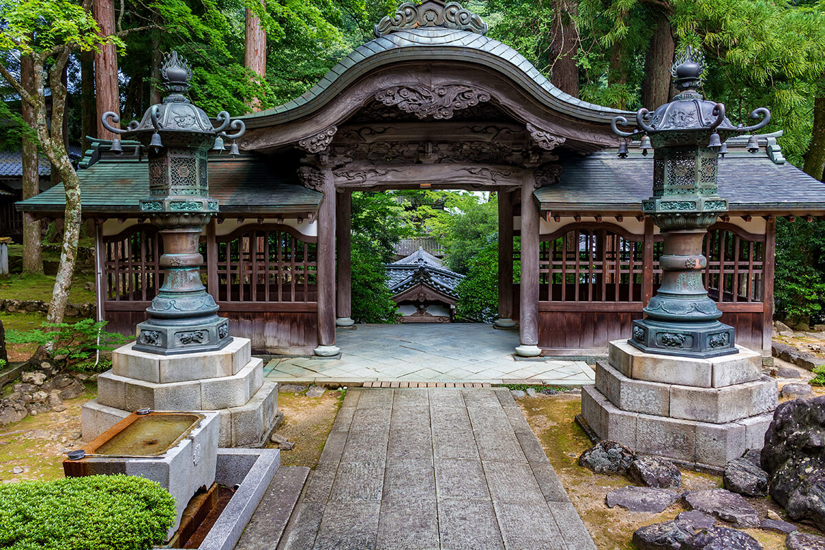 An image of a serene and peaceful Zen temple surrounded by lush greenery. The temple has a traditional Japanese architectural style with sloping roofs, wooden beams, and sliding doors. A stone pathway leads to the entrance of the temple. In the foreground, there is a small pond with a stone lantern. The overall atmosphere of the image is one of tranquility and spiritual contemplation.