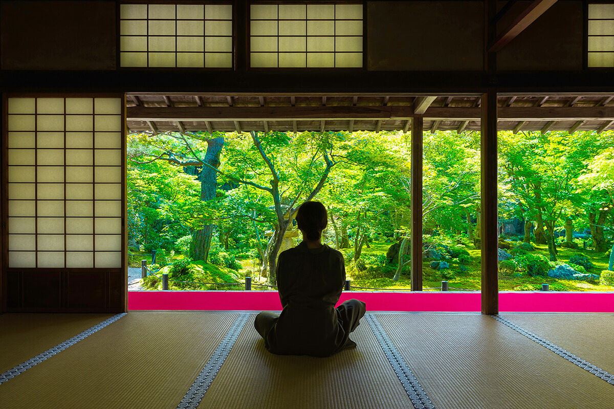 A person sitting in a traditional Japanese tatami room, surrounded by natural wood and paper walls. The person is sitting cross-legged on a cushion, with their eyes closed, in a state of deep meditation. The room is illuminated by soft, natural light filtering in through the paper-covered windows. 