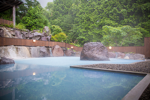 A hot spring (onsen) in Yufuin Tsukanoma, a wellness retreat in Japan. The onsen is surrounded by rocks and features a rectangular pool of hot, steamy water. The water is crystal clear and reflects the natural light, giving the space a warm and relaxing atmosphere. 