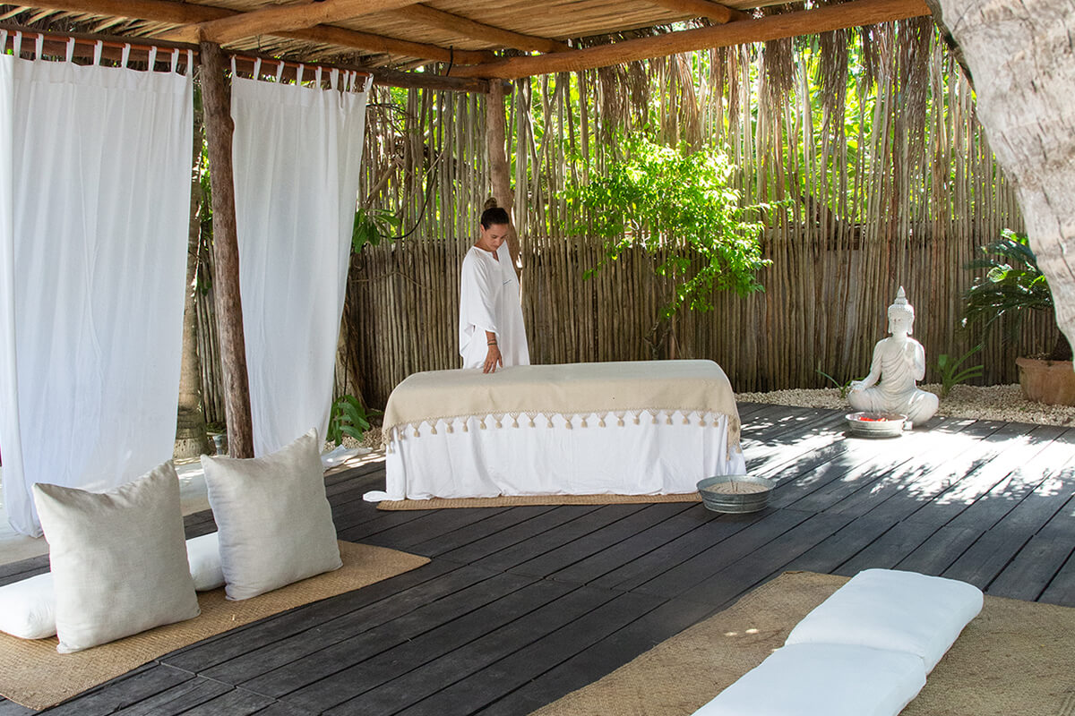 An outdoor setting with a wooden platform where a woman is standing over a massage table with white linens. Overall, the image conveys a sense of tranquility and relaxation, with a focus on holistic wellness practices such as yoga, meditation, and massage.