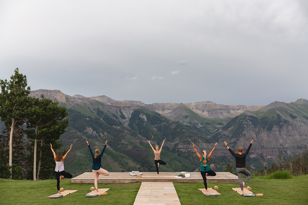 A yoga class in a natural outdoor setting. The class is being held on a grassy lawn, surrounded by mountains and trees. Several yoga mats are spread out on the grass, and a group of people are practicing yoga poses, with a teacher at the front of the class leading the session. 