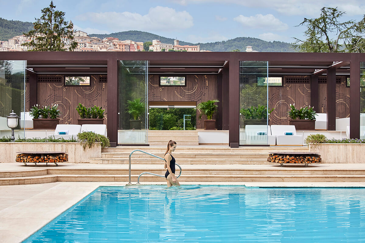 A woman stepping into an outdoor swimming pool at The Ranch Italy wellness retreat. The pool is surrounded by lush green trees and plants, with lounge chairs and umbrellas for relaxation. The water in the pool is a crystal clear blue, and the image captures the peaceful and serene atmosphere of the retreat's outdoor space.