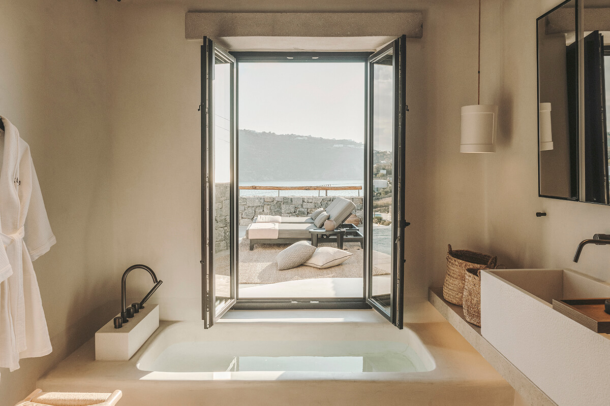 A luxurious bathroom in a hotel suite with a stunning view of the sea. In the background, a large glass window offers a breathtaking view of the sea, with the sun setting in the distance. 