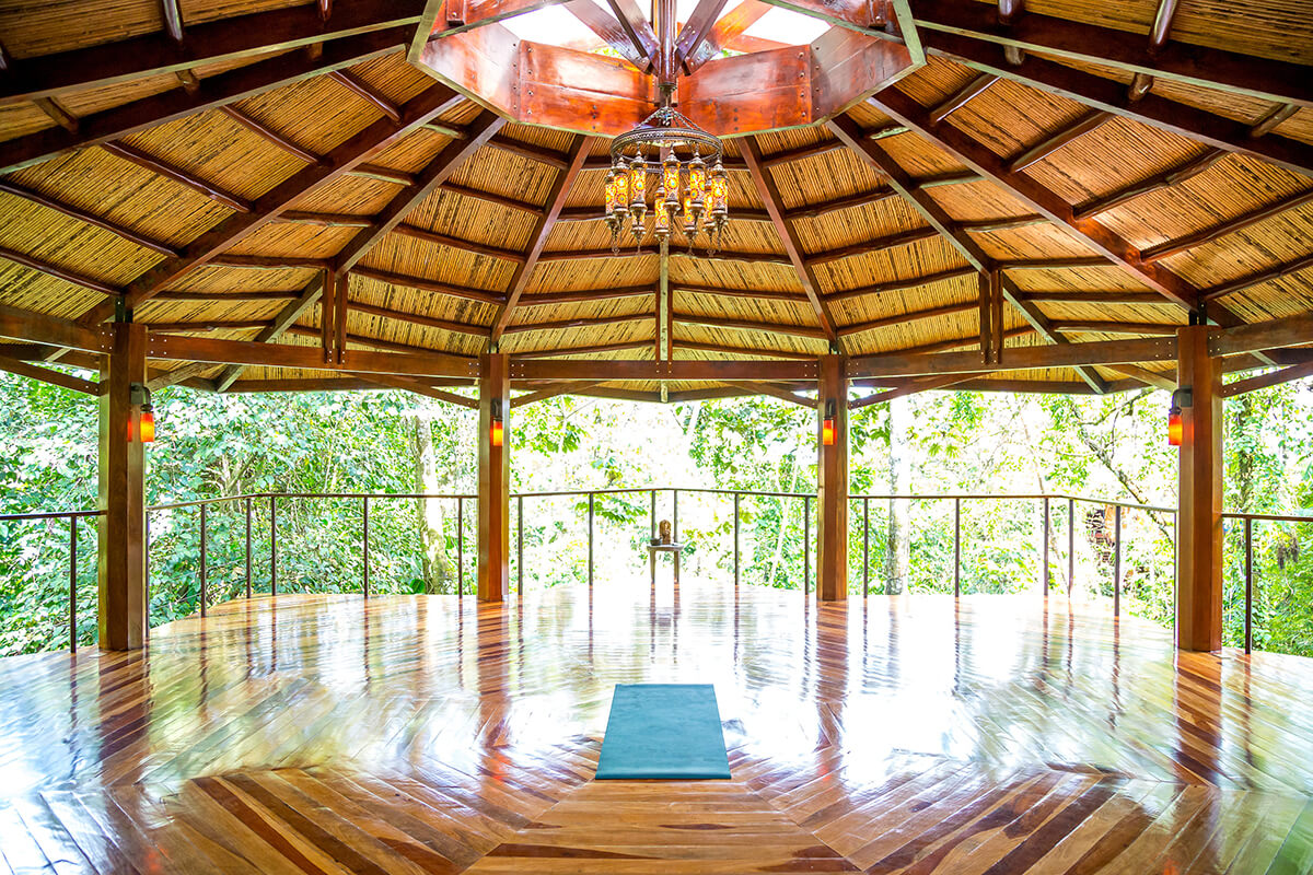 A serene outdoor space with a thatched-roof pavilion surrounded by lush tropical vegetation. The pavilion has wooden floors and pillars, and in the center of the pavilion, there is a wooden platform with yoga mats laid out. The background is filled with tall trees and dense greenery. The overall ambiance of the image is calm, peaceful, and harmonious with nature.