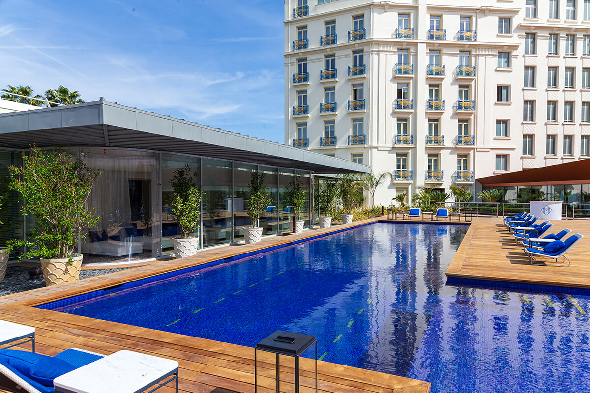 A luxurious outdoor swimming pool surrounded by several lounge chairs and umbrellas. The pool is a deep blue color. The area is well-manicured, with tall buildings in the background. The atmosphere is peaceful and relaxing, with the sunlight shining down on the pool area.