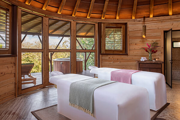 A luxurious spa room, with a serene and calming ambiance. There are comfortable white massage tables with a gray blanket, the room has large windows that let in natural light. 