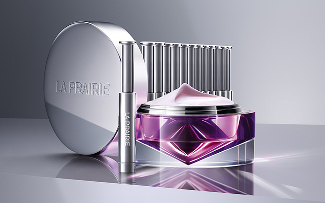 A purple bottle of La Prairie Anti-Aging Regenerating Cream next to a silver La Prairie Platinum Rare Haute-Rejuvenation Mask. Both containers sit on a light-colored table.