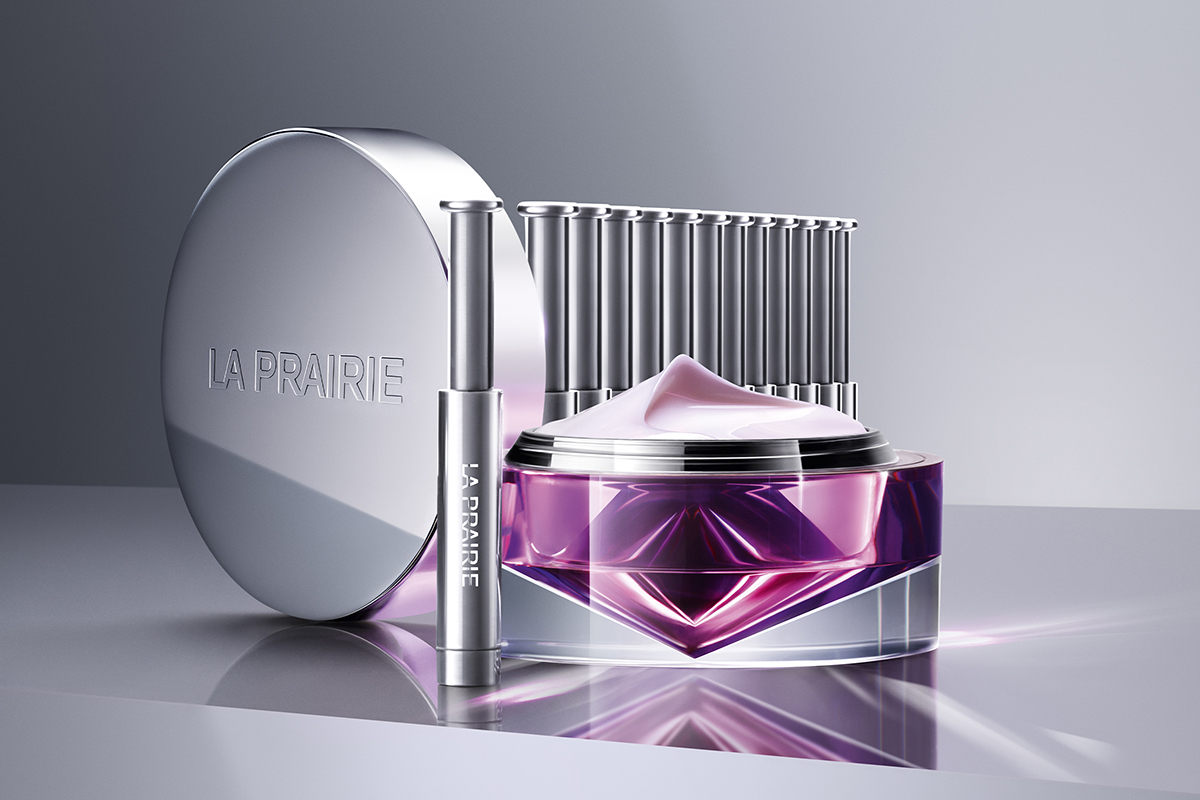 A purple bottle of La Prairie Anti-Aging Regenerating Cream next to a silver La Prairie Platinum Rare Haute-Rejuvenation Mask. Both containers sit on a light-colored table.