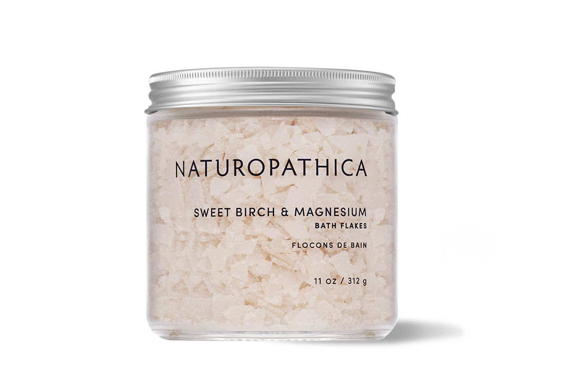 A glass jar of Naturopathica Sweet Birch and Magnesium Bath Flakes with a white label and a black lid.
