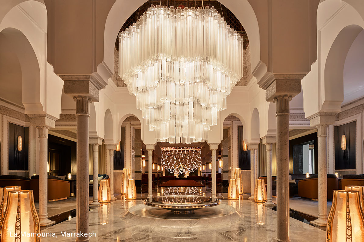 A large chandelier hangs from the ceiling of a grand hotel lobby. The chandelier is made of crystal and appears to have cascading tiers.