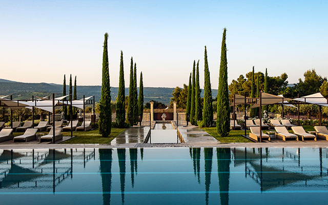 The resort swimming pool is surrounded by tall green trees and lounge chairs in Coquillade, Provence, France.