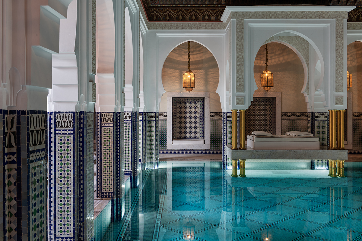 An indoor Moroccan-style swimming pool with arched entryways and white columns. In the center of the pool is a four-poster bed. The pool deck is tiled and there are two chaise lounges visible.