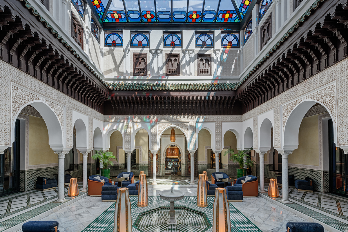 An ornate courtyard with a central fountain and a stained-glass ceiling. The fountain has multiple tiers and a cascading water feature. The stained-glass ceiling is colorful and geometric.