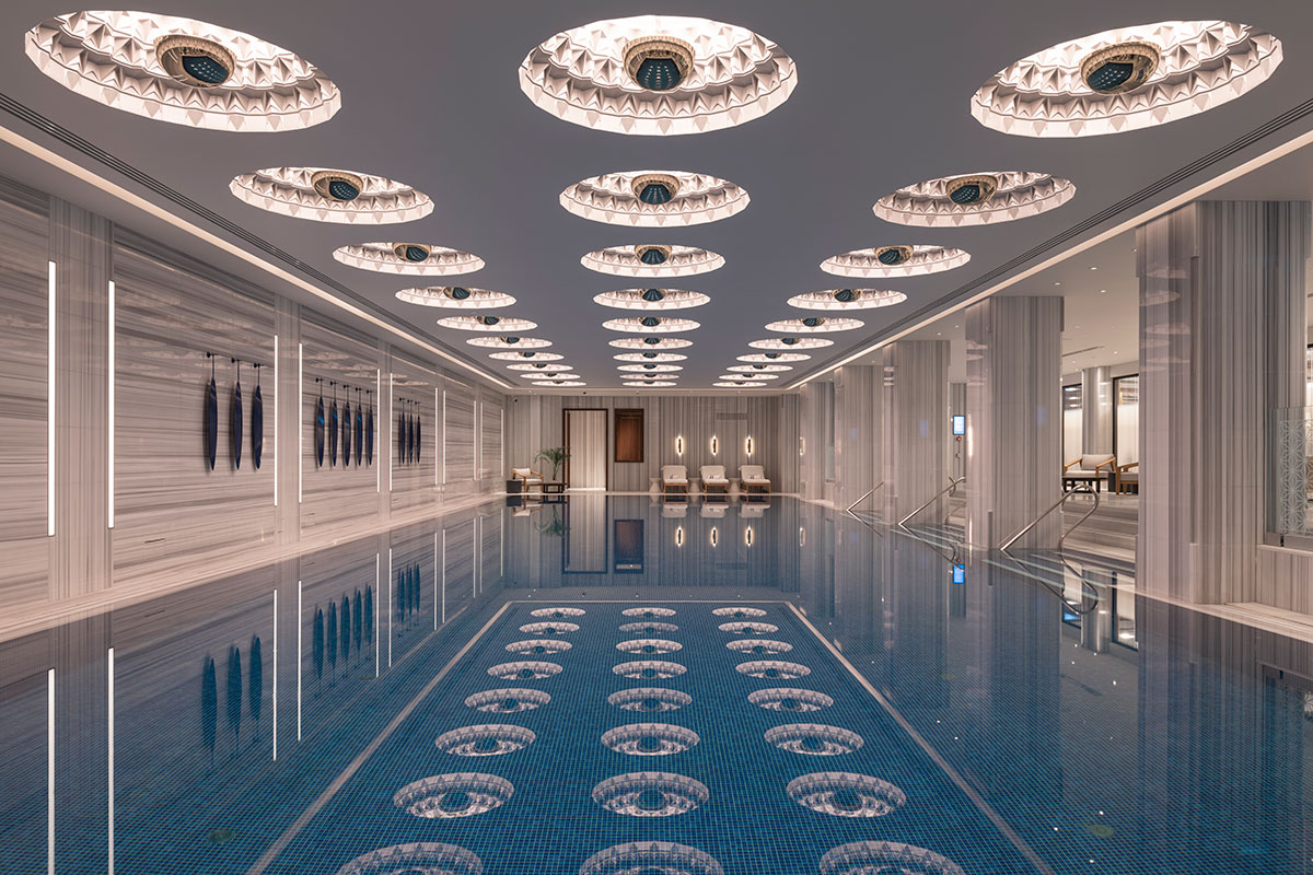 An indoor Moroccan-style swimming pool with arched entryways and white columns. In the center of the pool is a four-poster bed. The pool deck is tiled and there are two chaise lounges visible.