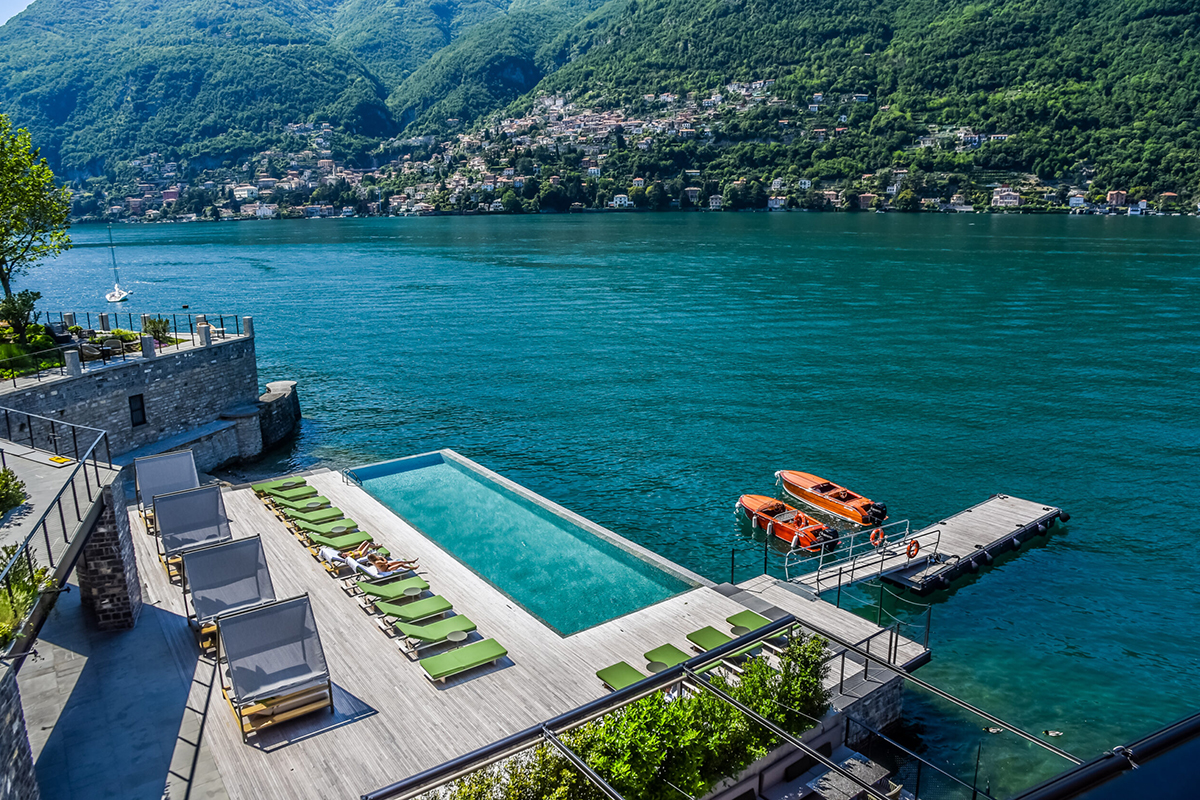 A rectangular infinity pool at the Il Sereno Hotel with a wooden deck juts out over Lake Como in Italy. In the distance are lush green mountains.