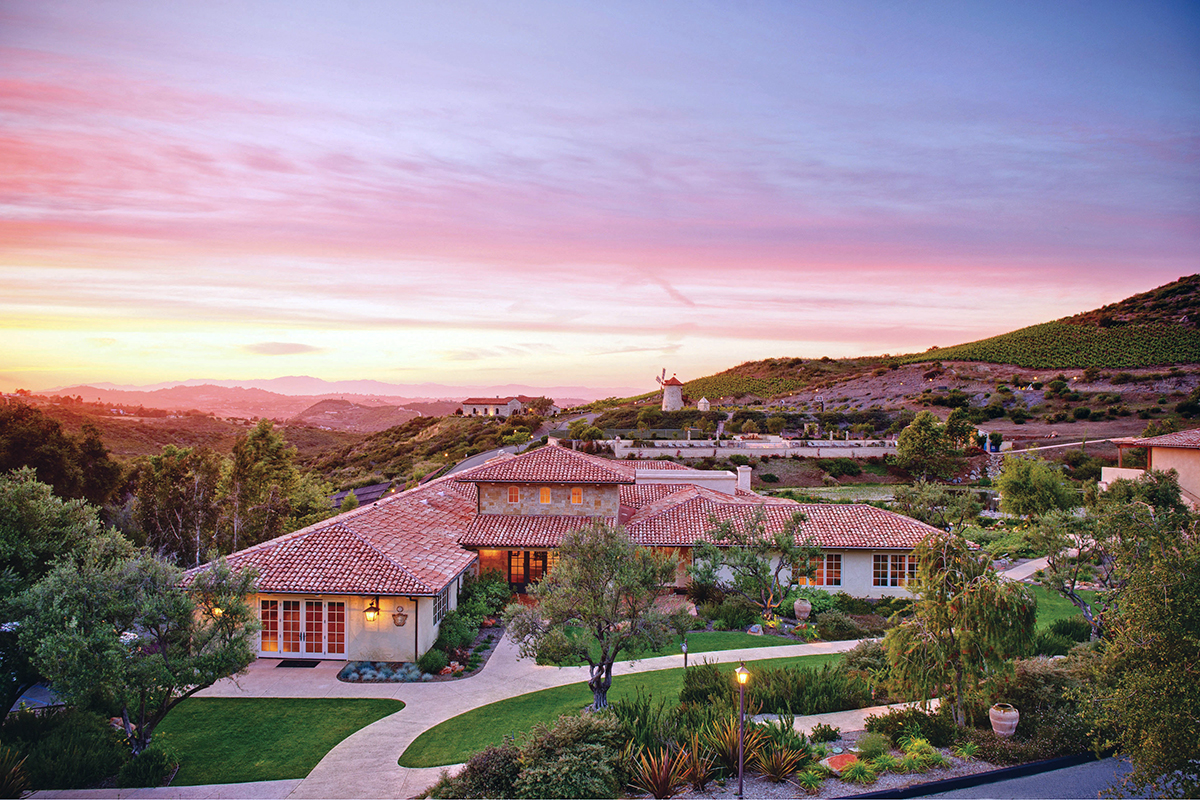 Large house with a red roof and windmill on a hill at sunset. The house is located at Cal-a-Vie Health Spa in Vista, California.