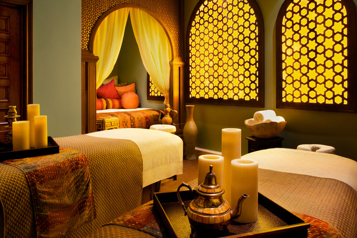 A pair of massage tables in the foreground covered in white linens in a warm-lit room with candles.