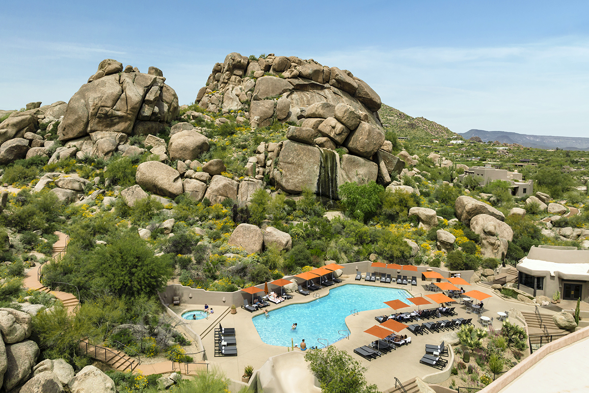 An outdoor pool area with lounge chairs surrounded by large natural rock formations with lush green trees.