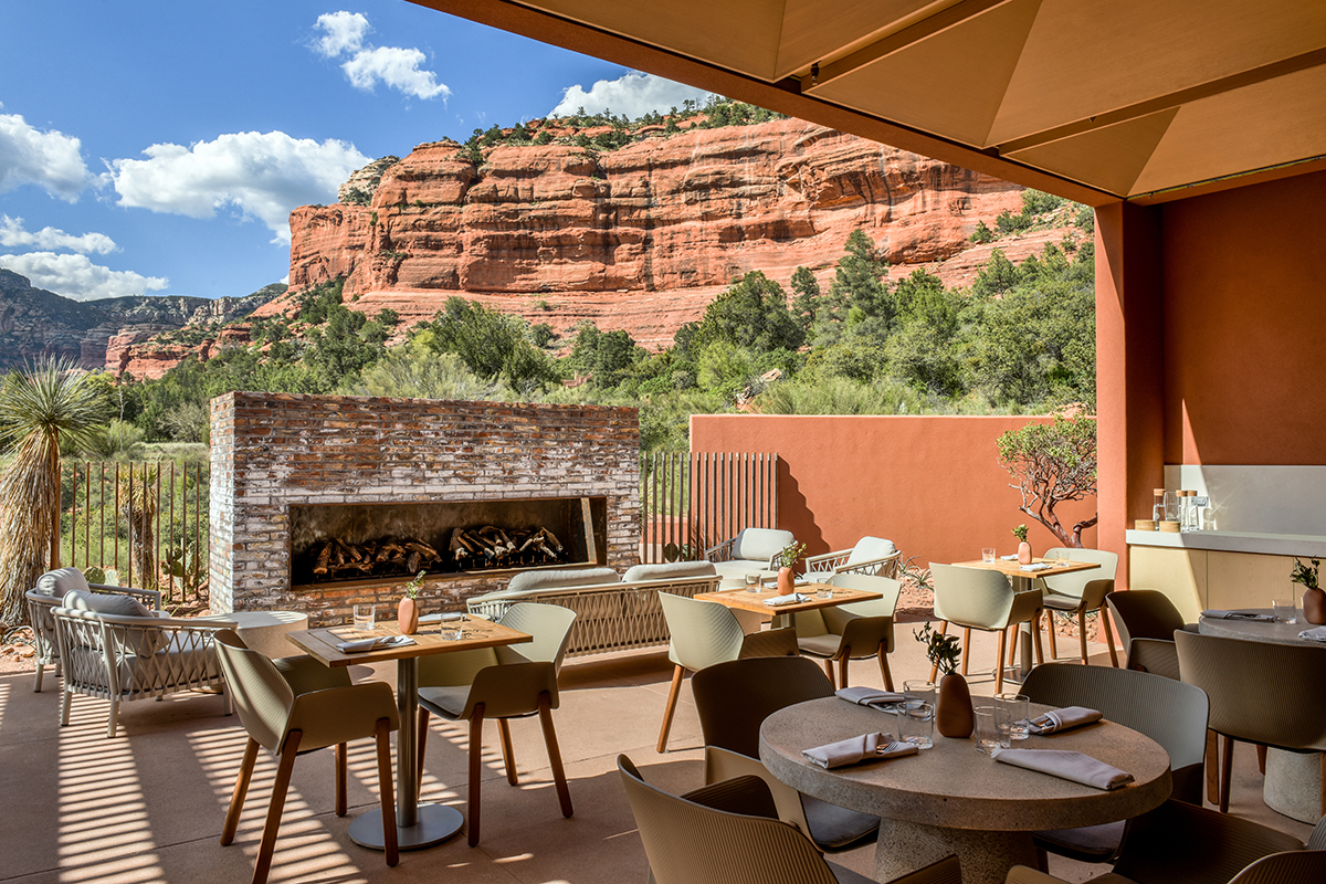 Stone patio with wooden tables, chairs, and a fireplace in front of a desert landscape.