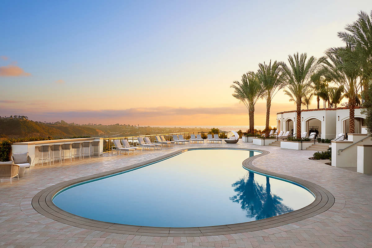 A large, curvy-shaped swimming pool surrounded by tall palm trees and a paved deck next to a red tile roof building.