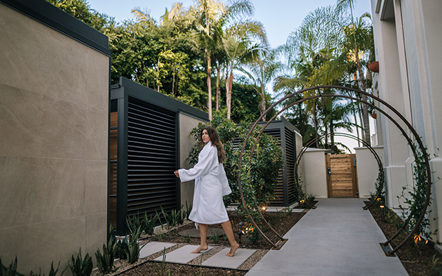 A woman in a bathrobe is about to enter a massage pod with rich wood accents enclosed in a walled tranquil environment with circular vine trellises.