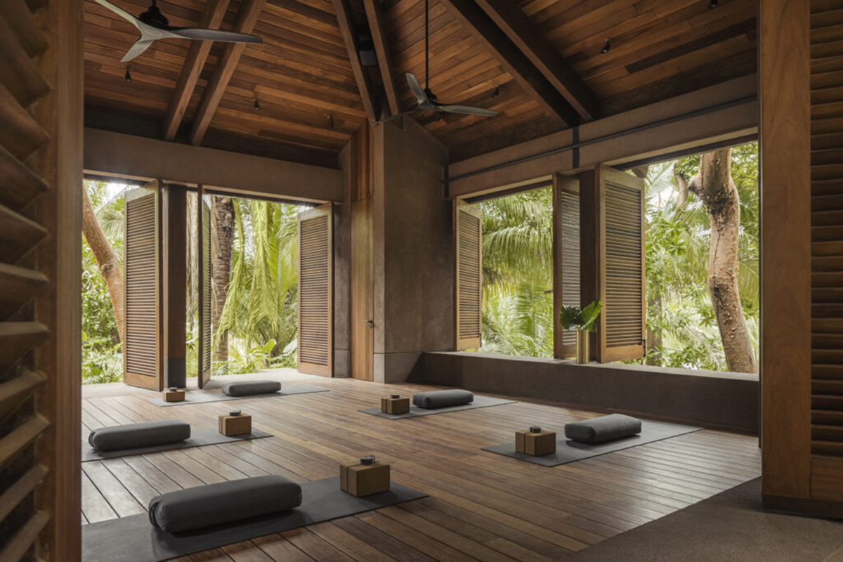 A spacious yoga studio with light brown walls and a wooden ceiling. Rolled-up yoga mats and meditation pillows are scattered on the wooden floor.