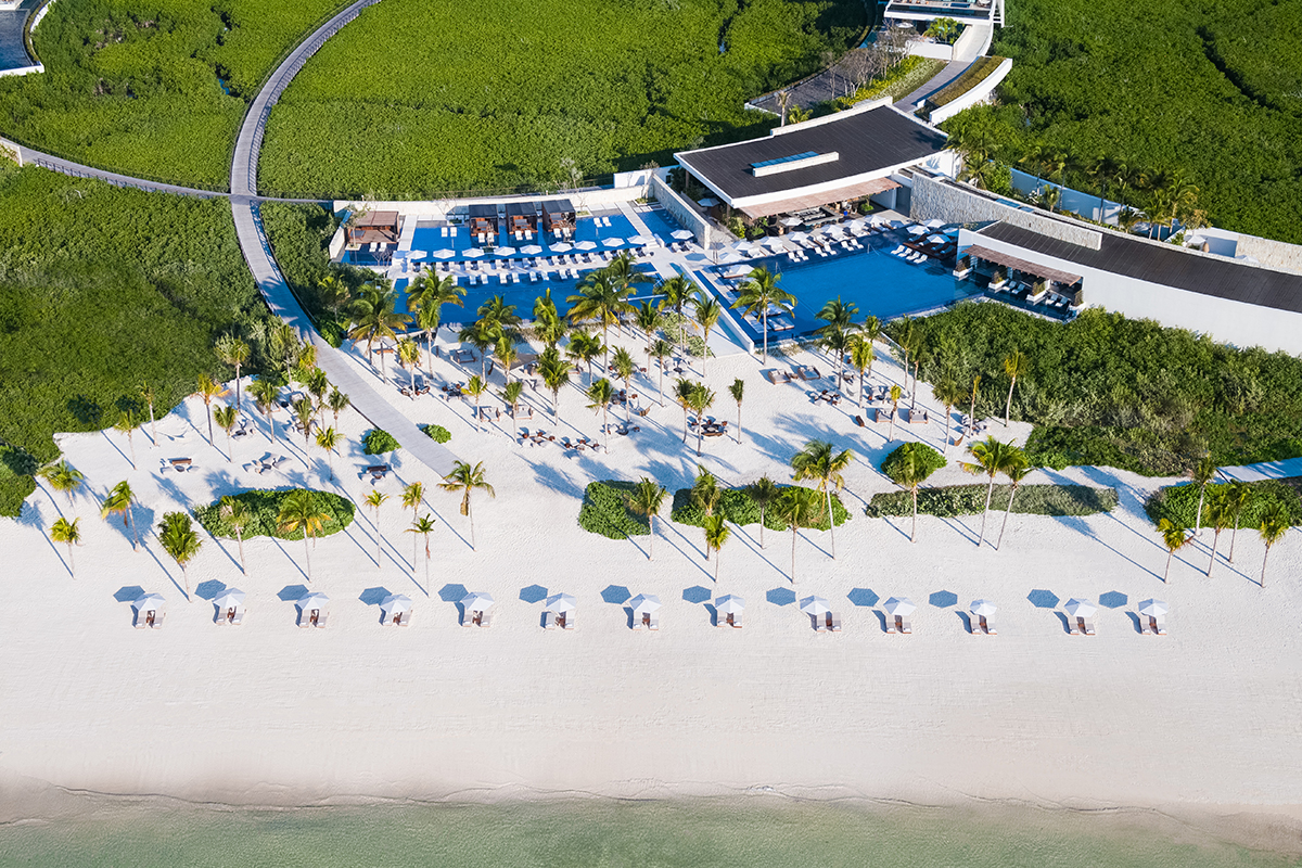 An aerial view of a beach resort with a rectangular swimming pool, white sandy beaches, palm trees, lounge chairs, and colorful umbrellas.