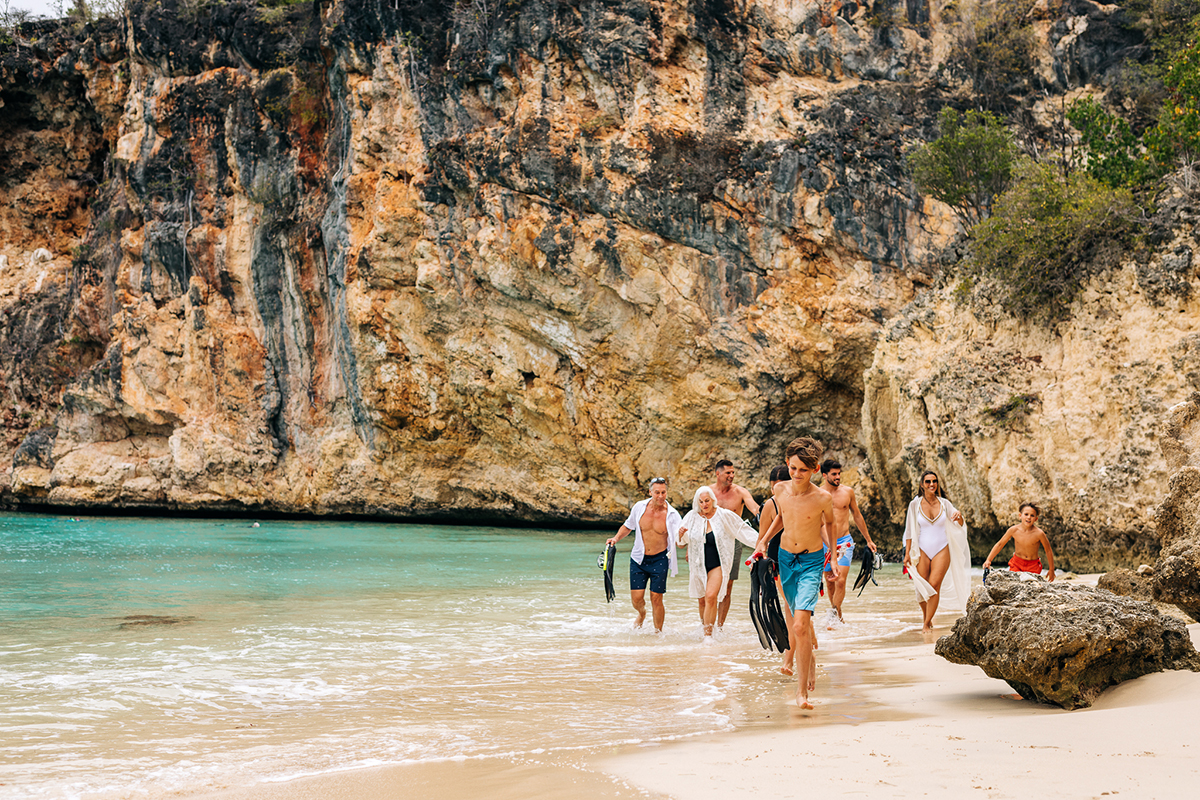A group of people walking along the ocean's edge on a secluded white sandy beach in their swimsuits, a few carrying scuba fins, in the background is a large cliff wall of multi-colored rock layers.
