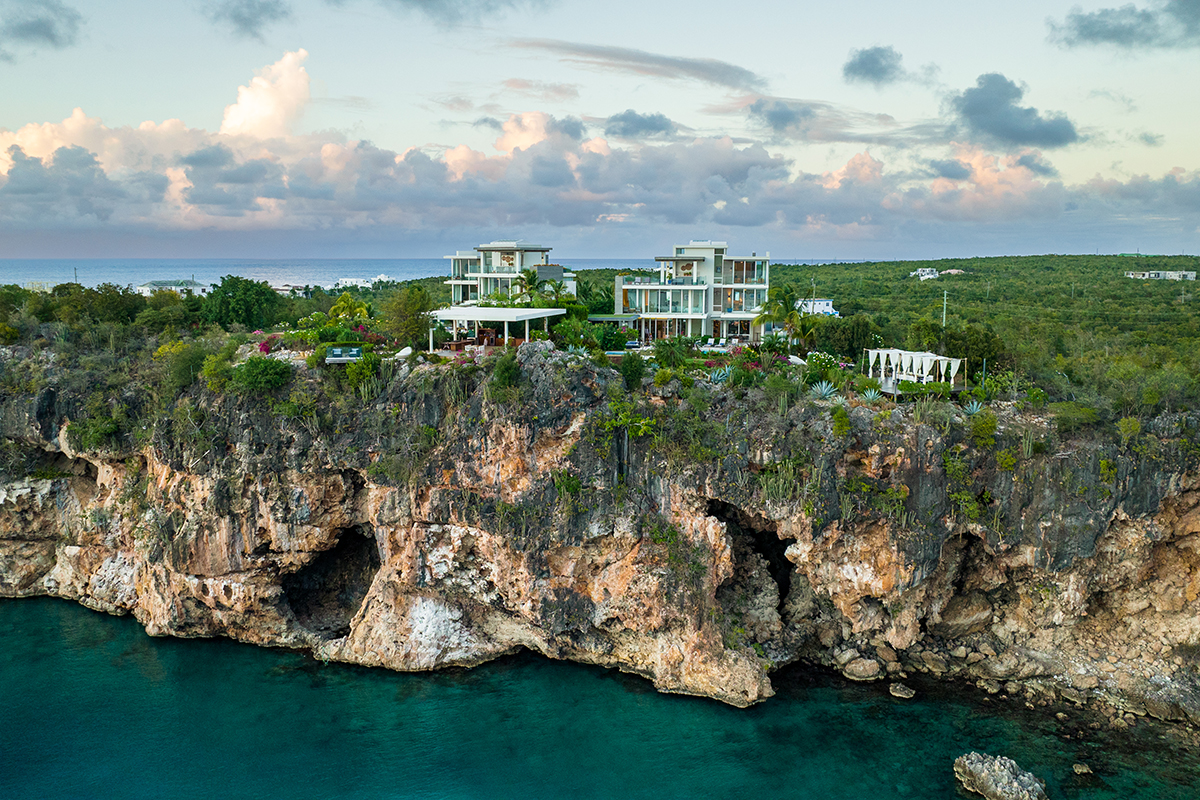 A modern house with a balcony, perched on a cliff overlooking the vast ocean. The ocean is a deep blue color and there are whitecaps on the waves. Lush green vegetation surrounds the house.