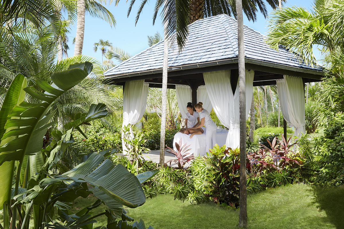 Outside underneath a hut with curtain walls, 2 massage therapists work on a resort guest in a lush beautiful palm garden setting.