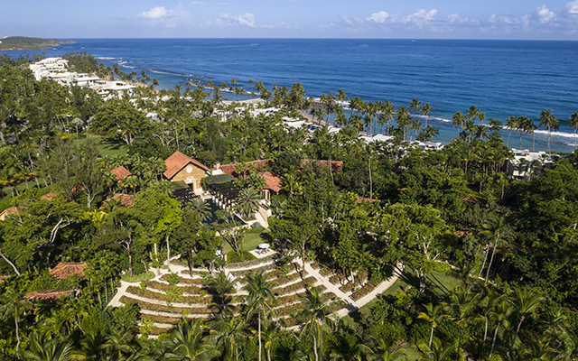 An aerial view of a tropical beach resort with white sand and turquoise water surrounded by lush green vegetation and palm trees.