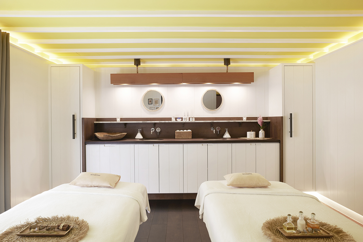 2 massage tables sit in the foreground covered in white linens with wooden trays resting on grass mats, in the background are white cabinets with rich dark wood countertops and two round mirrors hanging on the wall above it.