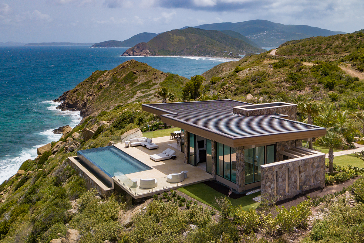A modern house with clean lines, built on top of a cliff overlooking a vast ocean. The house has a balcony with glass railings. The location is Oil Nut Bay.