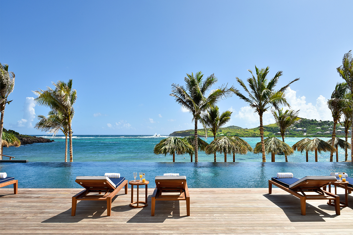 A luxurious infinity pool with a blue tile lining, overlooking a sparkling turquoise ocean. Tall palm trees with swaying fronds surround the pool deck. In the distance, a lush green mountain range can be seen.