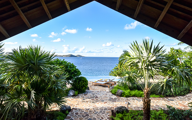A beautiful ocean view from a beach house with a rock walkway, green plants, and palm trees. In the distance is a white sandy beach, lounge chairs, a bright blue ocean, clouds, and a mountain range across the water.