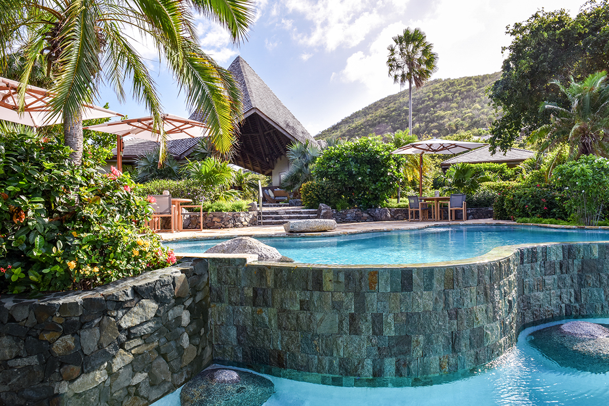 A large, curved elevated pool made out of chiseled rock blocks surrounded by a patio with umbrella tables and lush green ferns and palm trees.