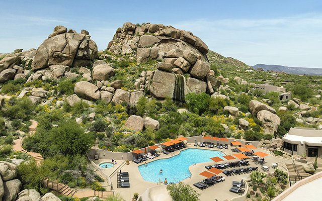 A large swimming pool with a waterfall cascading down rocks on one side. The pool is surrounded by lounge chairs and umbrellas on a sunny day.
